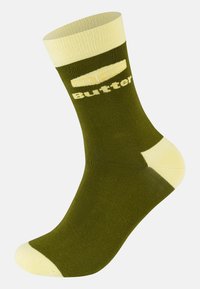 Olijfgroen sok met gele accenten aan de boord en de teen. Tekst "Butter" gedrukt aan de zijkant. Glad, rekbaar materiaal.