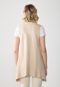 Beige ärmlös tunika med en avslappnad passform, tillverkad av mjukt tyg. Bakre delen har en rundad nederkant och inga synliga knäppningar eller detaljer.