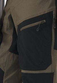 Close-up af brune og sorte udendørsbukser med en lynlåslomme med trækflapper og forstærkede sømme på stofpanelerne.