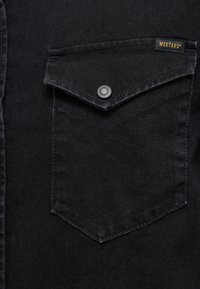 Szczegół czarnej koszuli z denimu z ostrą kieszenią na zatrzask i białym przeszyciem, metka z logo "MUSTANG."