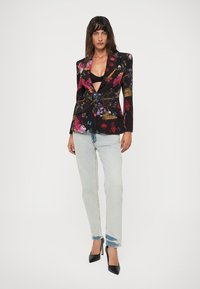 Donna che indossa un blazer floreale nero sopra una maglietta nera, jeans azzurri chiari e decolté nere a punta, in piedi contro uno sfondo bianco.