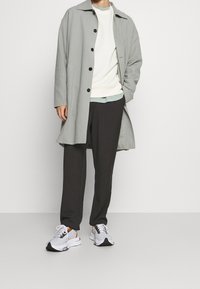 Manteau long gris clair avec boutons noirs, pull à col rond blanc, pantalon foncé, et baskets gris/blanc avec des accents orange.