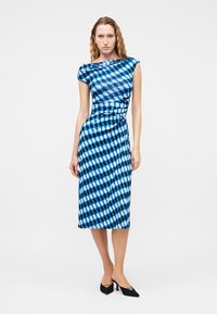 Γυναίκα που φοράει μπλε midi φόρεμα χωρίς μανίκια με μοτίβα και εφαρμοστή μέση, με μαύρες μυτερές γόβες, που στέκεται μπροστά σε απλό φόντο.