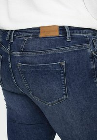 Tumši zila denim džinsi ar gludu tekstūru, ar ādas etiķeti uz jostas un tradicionālām aizmugurējām kabatām. Klasiskā piegriezuma dizains.