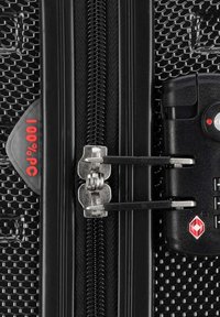 Close-up van een zwarte hardcase koffer met een gestructureerd oppervlak, dubbele ritsen en een TSA-goedgekeurd slot met een rode indicator die "100% PC" aangeeft.