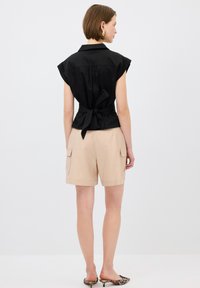 Camisa negra de manga corta con un lazo en la parte de atrás, combinada con pantalones cortos cargo beige. Pendientes de aro dorados notables y zapatos deslizables con estampado.