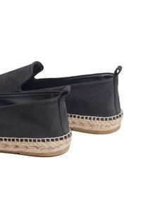 Scarpe slip-on in suede nero con suola in juta testurizzata e anelli laterali per una facile calzata, caratterizzate da un design semplice e colore neutro.