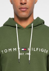 Zöld kapucnis pulóver pamutból, elöl található logóval, amelyen az olvasható: "TOMMY HILFIGER EST. 1985." Fehér húzózsinórokkal és ezüst fémlyukakkal rendelkezik.