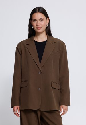 Donna con capelli lunghi e scuri indossa una giacca marrone oversize con top nero, orecchini argentati e anelli su sfondo neutro.