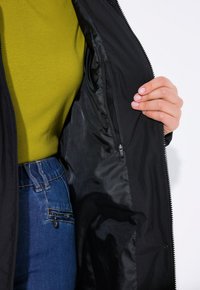 Interiér černé bundy ukazuje hladkou látku a obsahuje kapsu na zip. Model má na sobě zelený top a modré džíny.