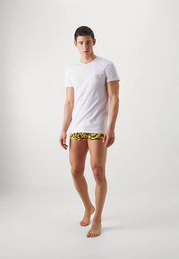 Νεαρός άνδρας που στέκεται ξυπόλητος, φορώντας ένα απλό λευκό t-shirt και κίτρινες εσώρουχες με σχέδιο, σε λευκό φόντο.