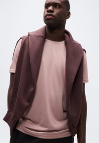 Νέος άνδρας που φοράει μωβ μπλουζάκι με ένα μπορντό φούτερ περασμένο στους ώμους του, κοιτάζοντας ελαφρώς προς το πλάι με φόντο ένα απλό τοίχο.