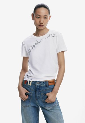 T-shirt en coton blanc avec le texte en cursive noir "Desigual" et "Fier d'être différent", associé à un jean en denim bleu clair avec une patch en cuir.