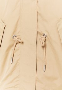 Beige jakke med en glat, mat finish. Har to snoretræk med knyttede ender og metaløjer. Simpelt design, ingen mønstre.