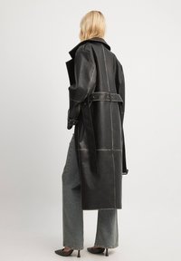 Schwarzer Ledertrenchcoat mit sichtbaren Nahtdetails, langen Ärmeln und einem taillierten Gürtel, getragen über hellblauen, weiten Jeans.