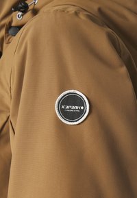 Manche de veste marron avec un patch rond noir et blanc cousu sur le haut du bras, portant l'inscription « Icepeak Established in 1996 ».