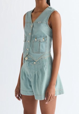 Kvinde iført lyseblå denim ærmeløs vest med knapper og matchende højtaljede shorts med plissering på ensfarvet baggrund.