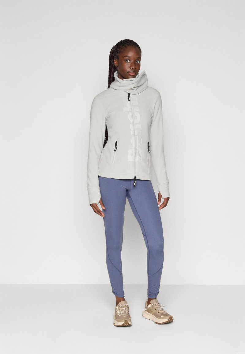 Lichtgrijze fleece vest met rits en hoge kraag, zwart logo en twee ritszakken, gecombineerd met blauwe legging en beige sneakers.