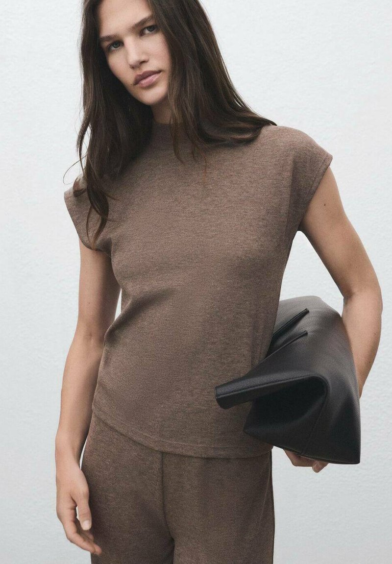 Maglione marrone lavorato a maglia con maniche corte abbinato a pantaloni coordinati; il modello tiene in mano una grande pochette in pelle nera con un design strutturato.
