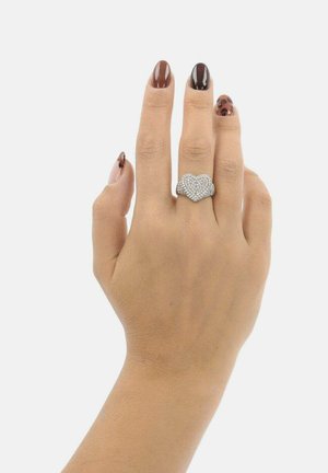 Hand met bruine en luipaardprintnagels die een zilveren hartvormige ring draagt, versierd met kleine edelstenen, aan de middelvinger.