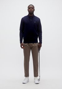 Homme portant un pull bleu marine, un pantalon beige, des baskets blanches, tenant un club de golf, debout devant un fond blanc uni.