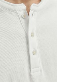 Acercamiento de una tela blanca texturizada con tres botones perlados en la tapeta de una camisa cerca del cuello.
