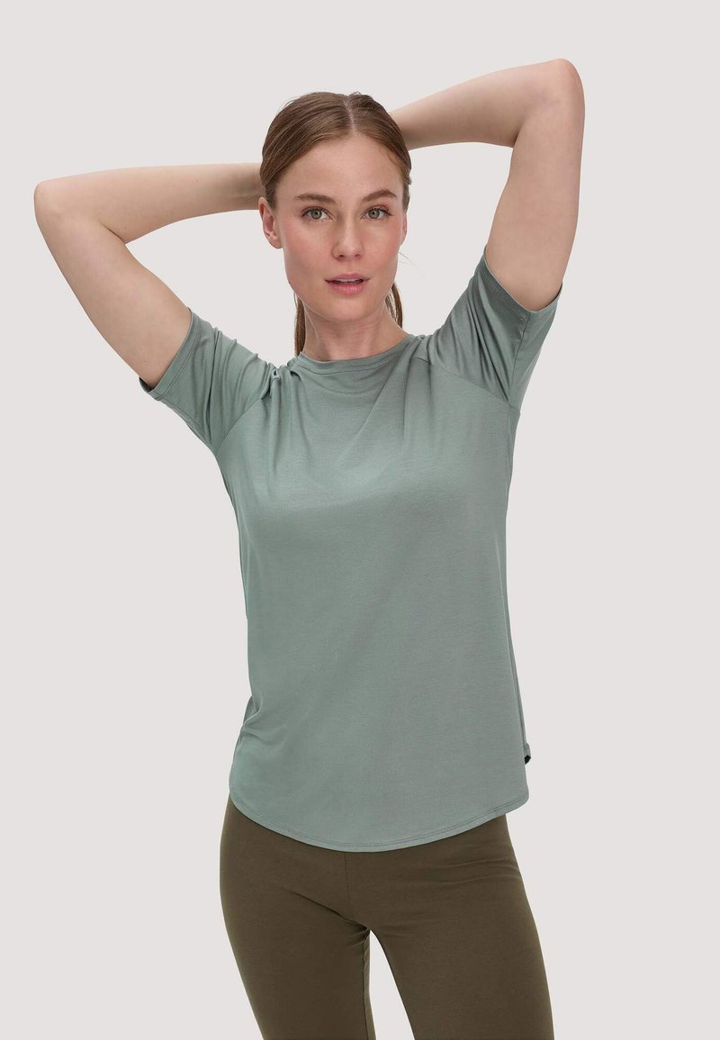Femme aux cheveux châtain clair attachés, portant un t-shirt vert sauge à manches courtes et un pantalon marron, les bras relevés derrière la tête.