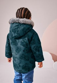 Teal puffjacka med grå fuskpälskant; har svarta mönster av byggfordon. Kombinerad med blå jeans, sedd bakifrån.