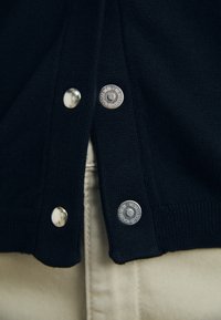 Granatowy sweter z dzianiny z metalowymi napami i ściągaczami na rękawach, noszony z jasną koszulą pod spodem.