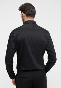 Zwarte geknoopte blouse gemaakt van gladde stof, met een klassieke kraag en lange mouwen. Het achteraanzicht toont een getailleerde pasvorm en een subtiele glans.