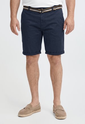 Un homme porte un short bleu marine à revers, une ceinture rayée, une chemise blanche et des mocassins beiges sur un fond uni.