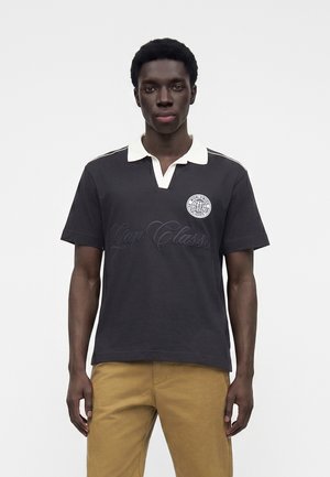 Ung mand, der står iført en sort polo med hvid krave og beige bukser mod en ensfarvet hvid baggrund.