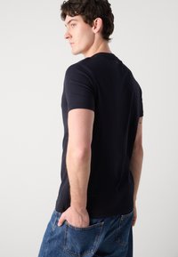 T-shirt marin à manches courtes en tissu doux. Avec un col rond et une coupe ajustée. Porté avec un jean en denim bleu, montrant la vue de dos.