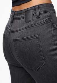 Černé džínové kalhoty s hladkou texturou, středním pasem a výraznými zadními kapsami. Detail s látkovou nášivkou nad pravou kapsou.