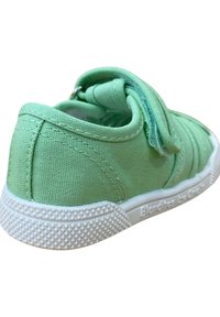 Zapatilla de lona verde con una correa de Velcro, parte superior texturizada, suela de goma blanca y detalle del logotipo en el lateral. La vista trasera resalta la costura.