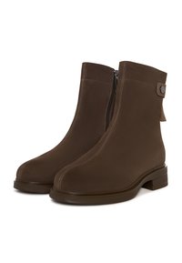 Un par de botas de tobillo de ante marrón con cremalleras laterales, punteras redondas, tacones bajos, costuras blancas y detalle de botón en un fondo blanco.
