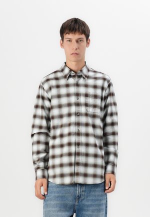 Young man wearing a long-sleeve, buttoned, black and white plaid shirt and blue jeans, standing against a plain white background.