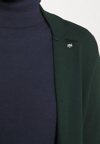 Green blazer with a notched lapel, paired with a dark navy turtleneck. A small silver bee pin accents the blazer's left lapel.