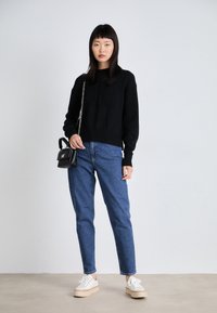 Maglione nero a maglia con orlo a costine, jeans blu a vita alta e sneakers bianche con plateau. Piccola borsa nera con tracolla a catena.