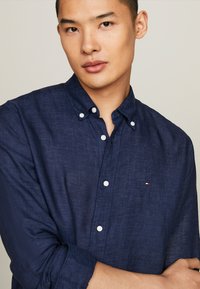 Camicia button-up blu scuro realizzata in tessuto testurizzato, con colletto classico e bottoni bianchi, e un piccolo logo ricamato sul petto.
