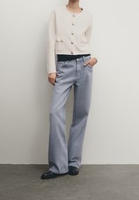 Light cream cropped jacket with gold buttons and pockets, paired with blue wide-leg jeans. The ensemble features a black top underneath.