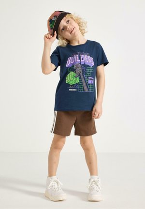 Lyshåret barn iført en Minecraft Builders navy T-shirt, brune shorts med hvide striber, hvide sneakers og holder en Minecraft kasket.