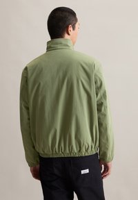 Lichtgroene, elastische taillejas met een hoge kraag en een gladde textuur; heeft een label op de zwarte broek. Gezichtspunt van achteren.