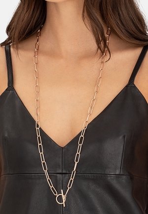 Femme portant un long collier en chaîne dorée avec de grands maillons rectangulaires sur un haut sans manches en cuir noir à col en V.