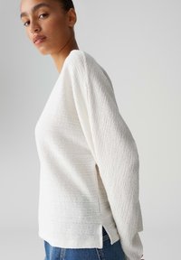 Giovane donna con maglione bianco a maniche lunghe dalla texture e jeans blu, in piedi contro uno sfondo grigio chiaro uniforme.