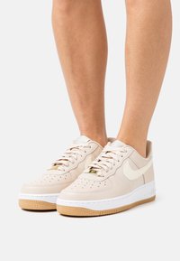 Beige leather sneakers with white sole and gum outsole. Features perforated toe, rounded shape, and contrasting cream swoosh. Gold accents on laces.