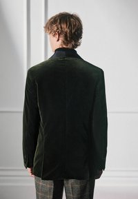 Ciemnozielona aksamitna marynarka z czarnym satynowym kołnierzem, z pojedynczym rozcięciem z tyłu i guzikami na rękawach, noszona na tle w kratę.