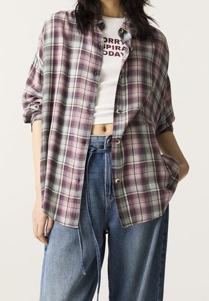Person wearing purple plaid button-up shirt over white cropped top with text, paired with loose blue denim jeans against plain background.