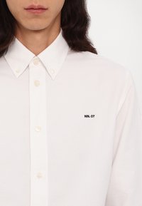 Personne portant une chemise blanche à boutons avec un col pointu et le texte noir "NN. 07" brodé sur le côté gauche de la poitrine.
