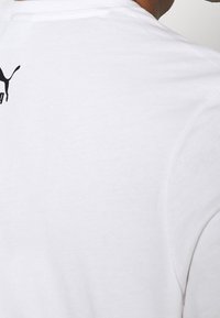 Bílá bavlněná tričko s kulatým výstřihem a krátkými rukávy, na zadní straně poblíž límečku se nachází malé černé logo Puma.